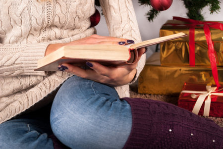 Girl in sweater holding book in hands and Christmas tree and gift box in background, close up,...