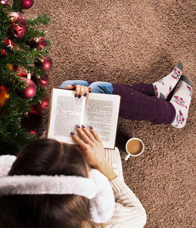 Female sitting on floor carpet and reading book next to Christmas tree and coffee cup, close up, top viewの写真素材