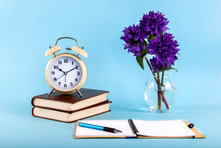 Open notebook with a pen, old clock on books and flowers in a vase on a blue background, blog...