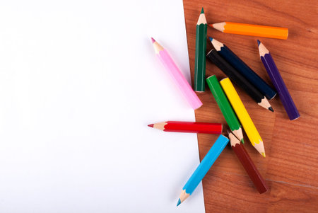 Colored wooden pencils spilled and white paper on table, close up