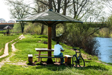 NIS, SERBIA - APRIL 05 2018: Alone man is sitting on bench and bicycle in park by the river on spring dayのeditorial素材