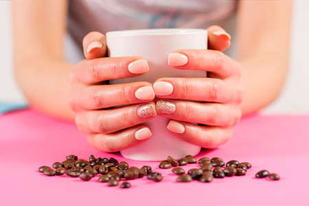 Young girl hand holding cup of coffee and coffee beans on pink desk. Manicure and Beauty and drink concept. Selective focus, close upの写真素材