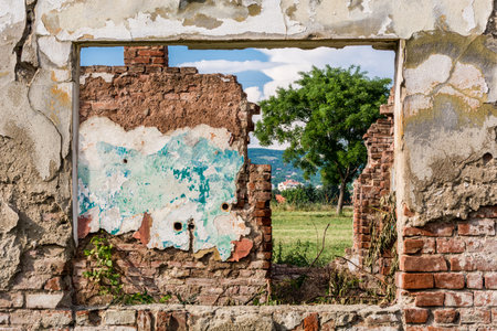 Damaged wall and window frame with view to ruins room and green field of grass, tree and the blue...