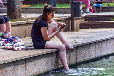 Niska Banja, Serbia - July 02, 2018 Young cute girl looking in phone and relaxing his legs in healthy spa water in tree shade. Vacation and enjoyment of the summer conceptのeditorial素材