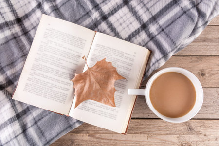 Open book with dry fallen leaf and cup of coffee on wooden background and retro blanket. Autumn...