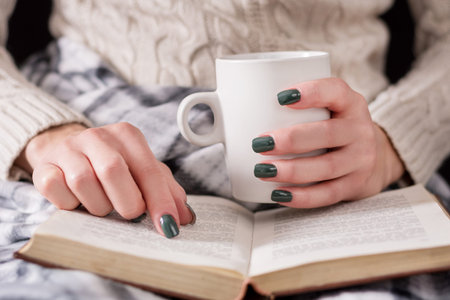 Woman reading book and holding cup of tea. Girl wears woolen sweater and covered with a retro...