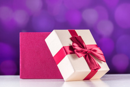 Red open gift box with bow on white desk and purple bokeh background. Christmas and holiday present...