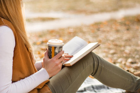 Young woman reading book in park and holding cup of coffee. Girl sitting on blanket on yellow fall leaf. Autumn concept. Close up, selective focusの写真素材
