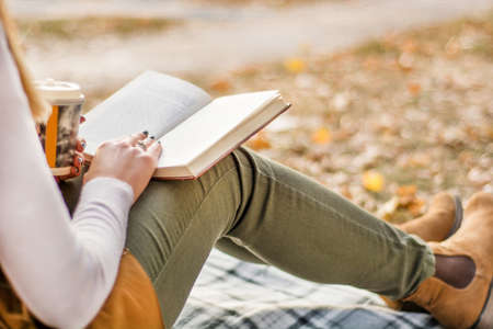 Woman with book on legs reading at retro blanket and blurred fall dry leaf in background on autumn day in park. Girl wears autumn clothes. Close up, selective focusの写真素材