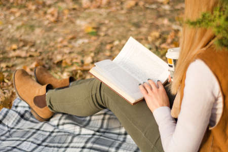 Young girl reading and holding book on legs at retro blanket at park on autumn day. Fall leaf blurred in background. Woman wears autumn clothes. Close up, selective focusの写真素材