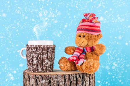 Hot tea cup in wool sweater with smoke and cute plush bear toy in winter cloths on wood log. It is snowing and blue background. Christmas holiday and winter idyll conceptの写真素材