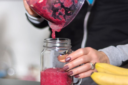 Girl pours red berry smoothie from blender to jar glass in kitchen at home. Healthy and detox food...