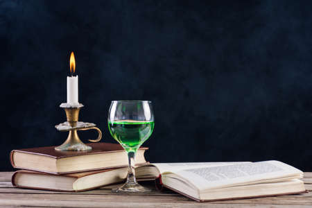 Absinthe drink in glass and books with candle in candlestick burning on retro wooden desk and smoky dark background. Green fairy inspiration and muse of writers and artistsの写真素材
