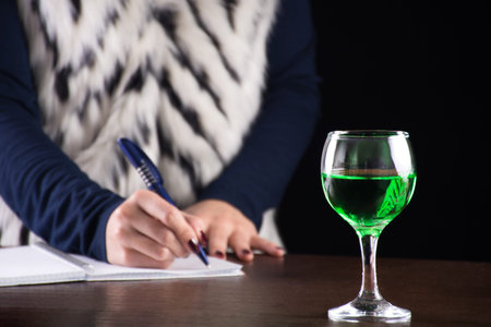 Absinthe green drink in glass and woman writing story notebook in background on old retro wooden...
