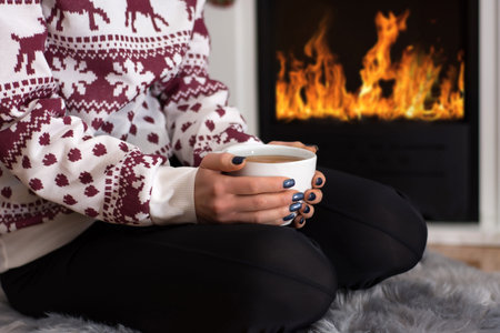 Girl drinking hot tea in front of the fireplace and sitting on furry carpet at home. Woman wears...
