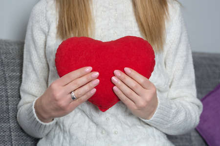 Young girl holding big red heart pillow in hand on her chest at home. Girl in white sweater. Woman day and Valentines day concept. Close up, selective focusの写真素材