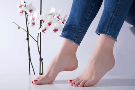 Female feet with red pedicure and jeans on legs, white orchid flowers in background. Pedicure and foot care concept in beauty studio. Close up, selective focusの写真素材