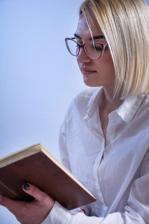 Blonde student girl reading book with glasses on eyes. Young women wears white elegant shirt. White...