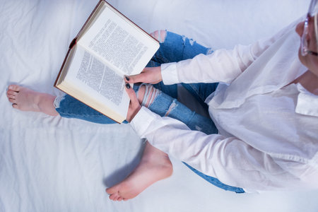 Student girl reading book in cozy bed and holding book on legs with bare feet. Relaxation and education concept. Close up, selective focusの写真素材