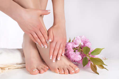 Female legs with bare feet and hands with french manicure and pedicure on white towel in spa salon and decorative pink flower in background. Nails polish concept. Close up, selective focusの写真素材
