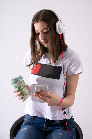 Teen girl listens to music on headphone and writes message on cell phone and drinking coffee...