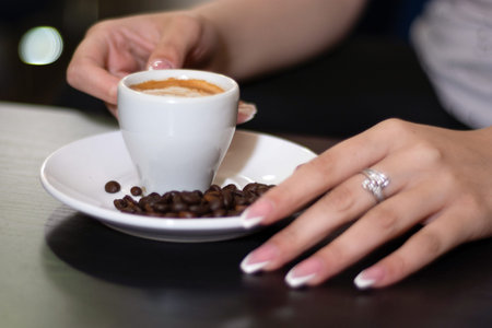 Girl hand holding espresso coffee cup on the desk in a bar. On saucer is blurred roasted coffee...