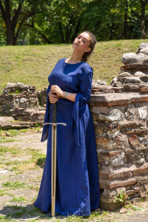 Nis, Serbia - June 15. 2019: Portrait of a medieval lady in a blue dress holding a sword in her hands in front of a medieval ruins building on a sunny day. Close up, selective focusのeditorial素材