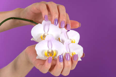Young woman hand with a lilac color nails polish gel and beautiful orchid flower decoration on purple background in the studio. Manicure and beauty concept. Close up, selective focusの写真素材