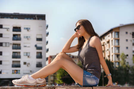 Girl sitting on a bench in a park and sunbathe legs on the sun. Young girl in shorts and sunglasses on the head on sunny summer hot day. Relaxation and summer concept. Close up, selective focusの写真素材