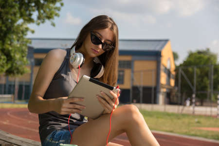 Young girl reading an electronic book on a digital tablet on a bench in the park on a sunny summer day in the sunset. Girl wears sunglasses and headphones on the neck. Education conceptの写真素材