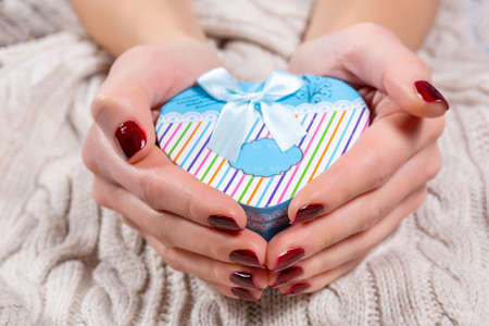 Girl hands holding a blue gift box in heart shape with a tie bow on woolen sweater background. Female hands with brown nails polish on fingers. Christmas or Valentines day holiday conceptの写真素材