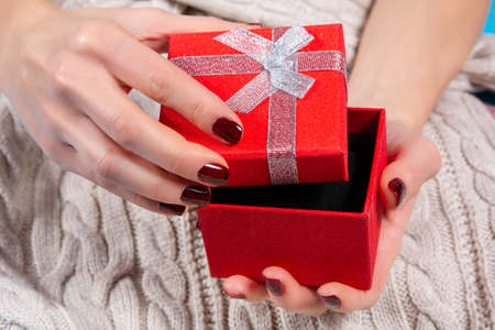 Girl hands holding and opening red gift box with a tie bow on woolen sweater background. Female hands with brown nails polish on fingers. Christmas or Valentines day holiday conceptの写真素材