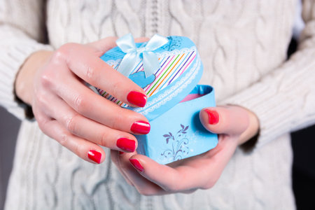 Girl hands holding and opening a blue gift box in heart shape and woolen sweater in background. Female hands with red nails polish on fingers. Christmas or Valentines day holiday conceptの写真素材