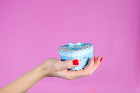 Woman hand holding a blue gift box in heart shape isolated on pink background in studio. Female hands with red nails polish on fingers. Christmas or Valentines day holiday and minimalism conceptの写真素材