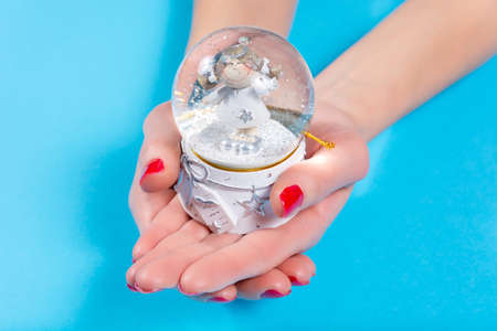 Woman hand holding ornament ball with snow isolated on blue background in studio. Female hands with red nails polish on fingers. Christmas or Valentines day holiday and minimalism conceptの写真素材
