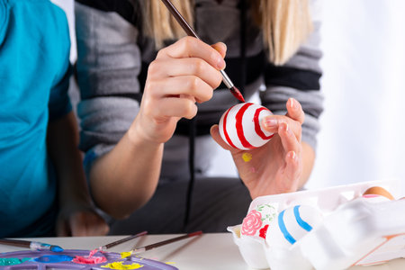 Female hand painting Easter eggs with a paintbrush on a table at home. The woman decorates the egg...