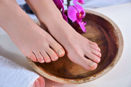 Female bare feet on wooden bowl with water and orchid flower and stone in beauty studio. Girl foot with natural color. Pedicure and relaxation foot concept. Close up, selective focus, top viewの写真素材