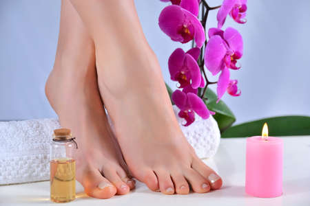 Women feet with natural nails polish color on white desk in the studio. Female legs on a towel with purple orchid flowers and candles in spa salon. Pedicure concept. Close up, selective focusの写真素材