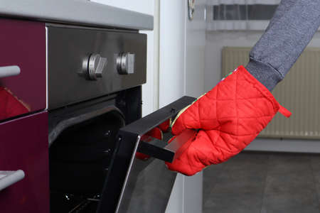 A woman hand with red heat protects kitchen gloves opening the oven. Food and prepare food in the kitchen concept. Close up, selective focusの写真素材
