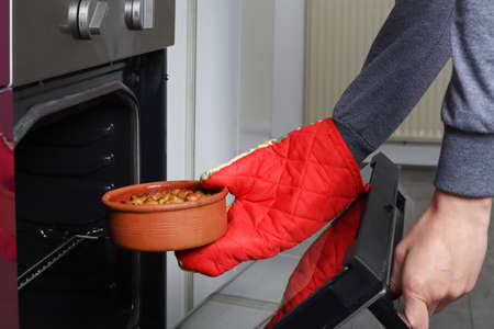 Woman hand takes it out terracotta saucepan with beans in the oven to bake at kitchen. Girl hand with red protect kitchen gloves. Traditional breakfast dish of beans. Close up, selective focusの写真素材