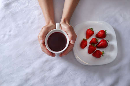 Woman hand holding a cup of tea next to a plate full of strawberries fruit. A plate and girl hands on a white tablecloth. Seasonal food and fresh fruit concept. Close up, selective focusの写真素材