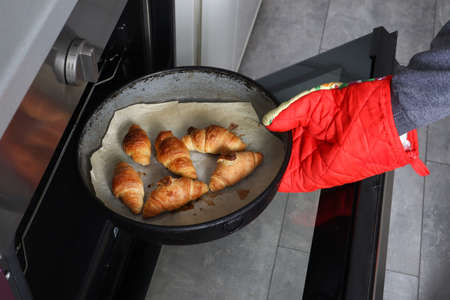 Girl hand with red protective glove takes out casserole dish with baked rolls from the oven in the kitchen at home. The process of preparing homemade rolls pastry. Close up, selective focusの写真素材