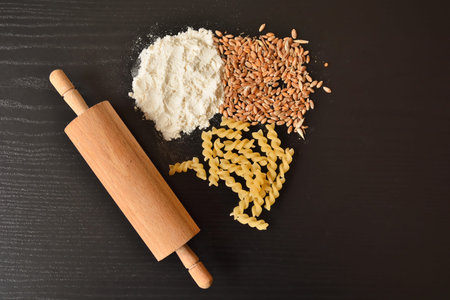 White flour, wheat grains, raw macaroni or pasta, and rolling pin on wooden black desk with copy...