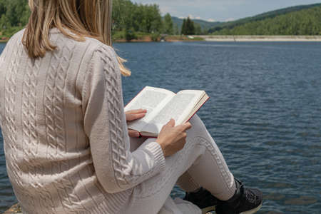 Girl reading a book near to lake water in a sweater on a sunny day. Blonde girl sitting on a large rock on the shore of the lake with a book on legs. Relaxation concept. Close up, selective focusの写真素材
