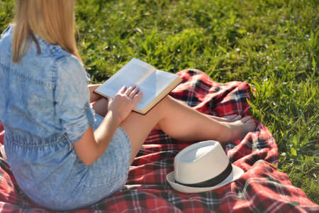Blonde girl holding book on legs and reading on sunny summer day in the park. Girl sitting on a red blanket in the green grass of a meadow. Relaxation in nature concept. Close up, selective focusの写真素材