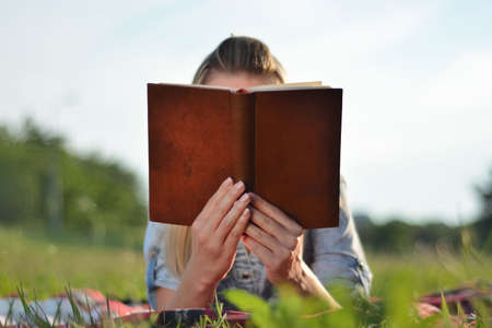 Girl reading book in nature on a sunny summer day. Girl holding opened a book and lying on her stomach on a blanket in the field. Relaxation in nature concept. Close up, selective focusの写真素材