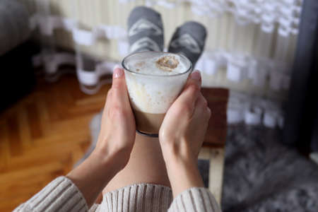Girl hands holding on legs glass cup of coffee with big milk foam at home in the living room. Hot drink conceptの写真素材