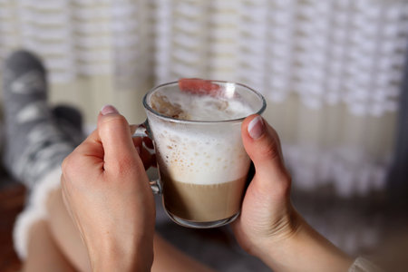 Girl hands holding glass cup of coffee with big milk foam on legs at home in the living room. Hot...
