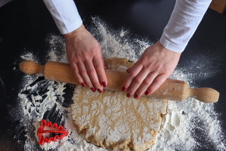 Female hands with a wooden rolling pin roll out the cookie dough on the kitchen table covered with...