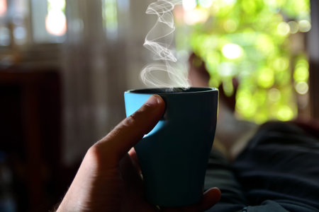 Blue cup of coffee in hand of man in bed at home. Man lying and relaxing on a hot summer day. Green...